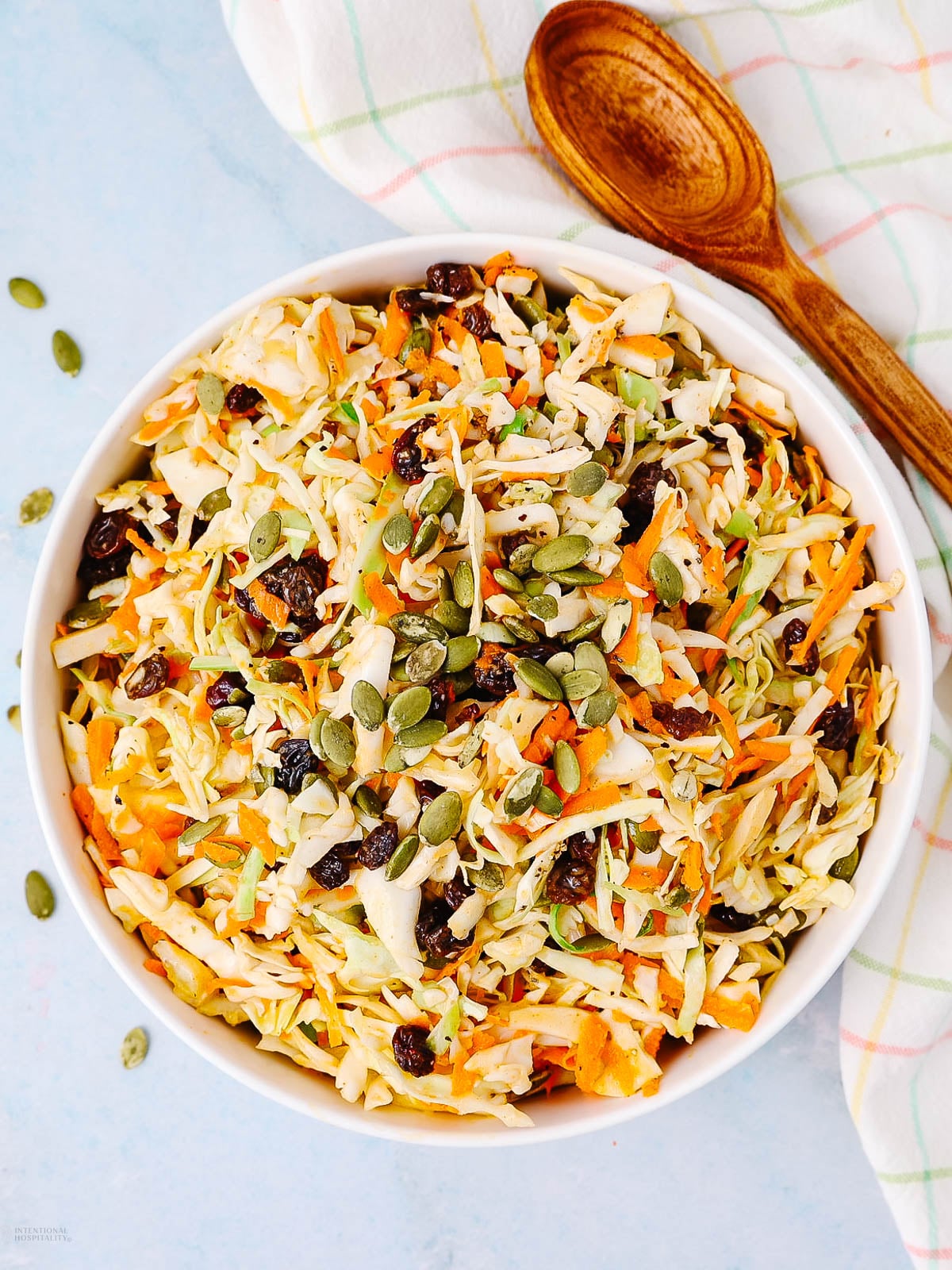 A bowl of coleslaw made with shredded cabbage, carrots, raisins, and pumpkin seeds sits on a light blue surface next to a wooden spoon and a checkered kitchen towel.