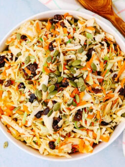 A bowl of shredded cabbage and carrot salad topped with raisins and pumpkin seeds, with a wooden spoon and a checkered cloth beside it on a light blue surface.