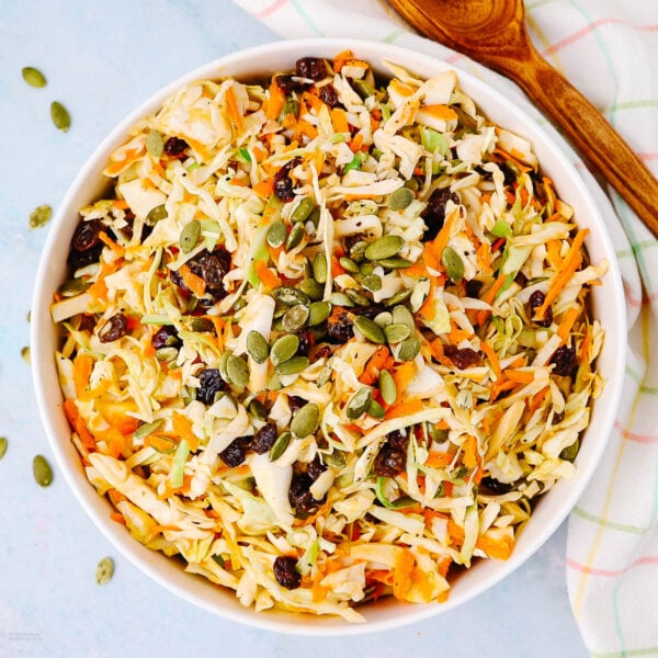 A bowl of shredded cabbage and carrot salad topped with raisins and pumpkin seeds, with a wooden spoon and a checkered cloth beside it on a light blue surface.