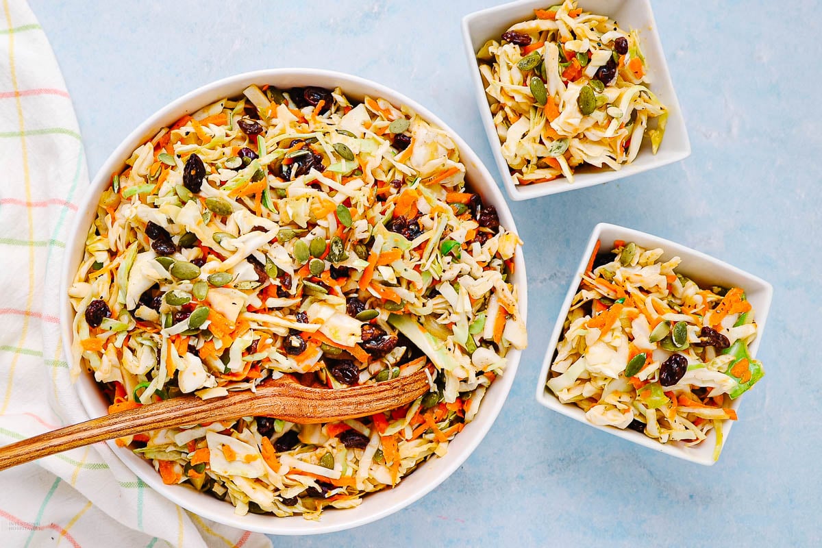 A large bowl and two smaller bowls filled with coleslaw made of shredded cabbage, carrots, raisins, and pumpkin seeds, with a wooden spoon resting in the large bowl, on a light blue surface.