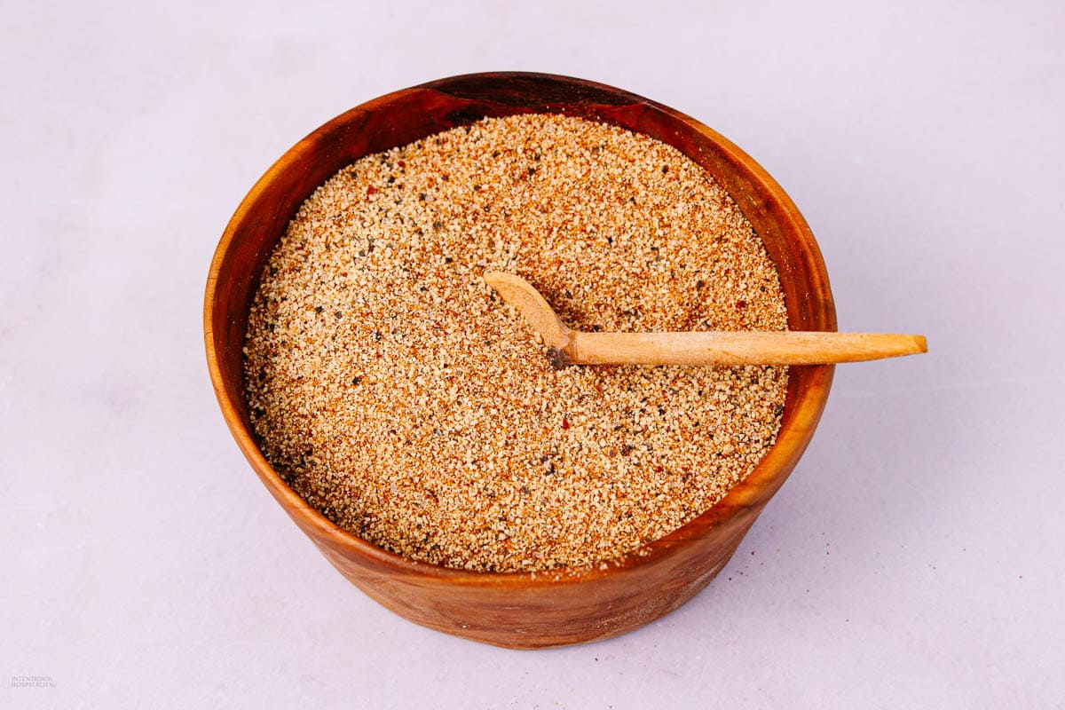 A wooden bowl filled with a coarse, mixed seasoning blend, accompanied by a small wooden spoon resting inside the bowl. The background is a plain, light surface.