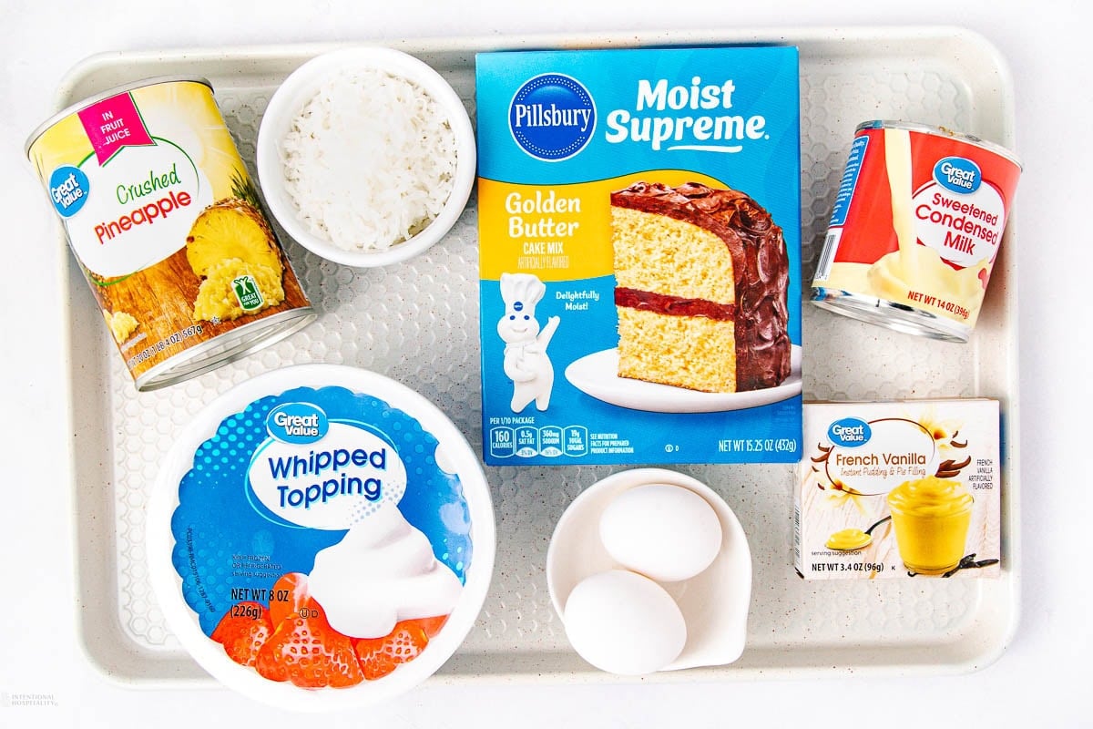 A tray with ingredients for a cake: a can of crushed pineapple, a bowl of shredded coconut, a box of golden butter cake mix, a can of sweetened condensed milk, whipped topping, two eggs, and a box of French vanilla pudding mix.