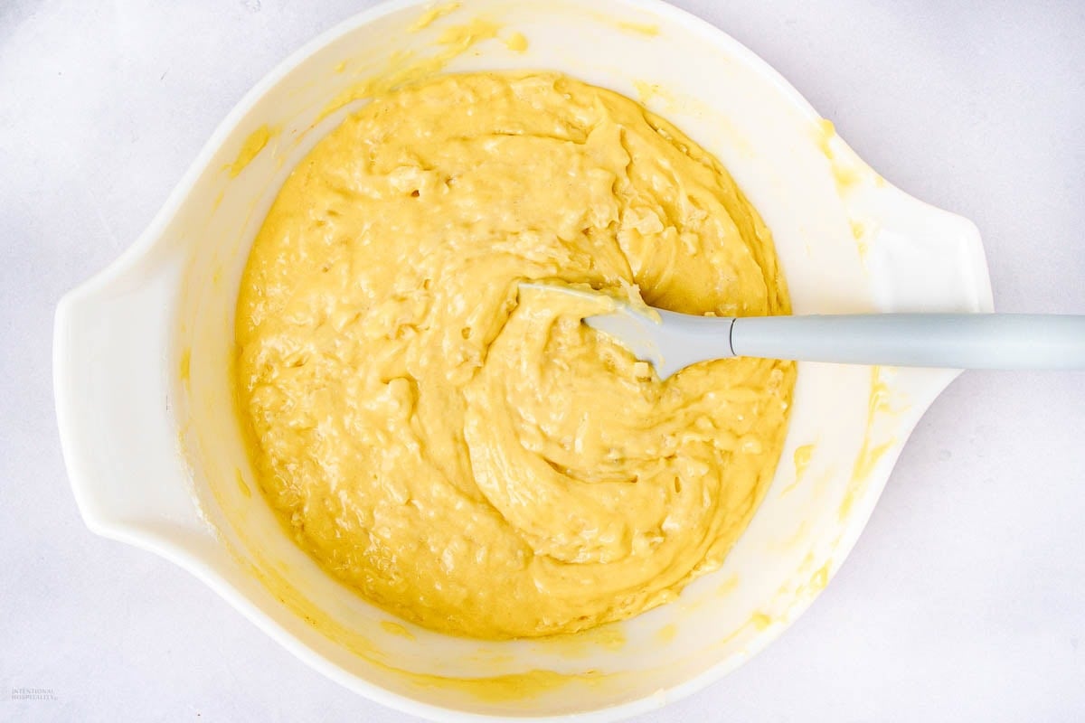 A white mixing bowl filled with yellow cake batter and a gray spatula resting inside, on a light surface.
