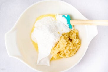 A white mixing bowl contains crushed pineapple, flour, sugar, and a creamy white ingredient, with a teal spatula resting on the side. The ingredients are unmixed and arranged in separate piles.