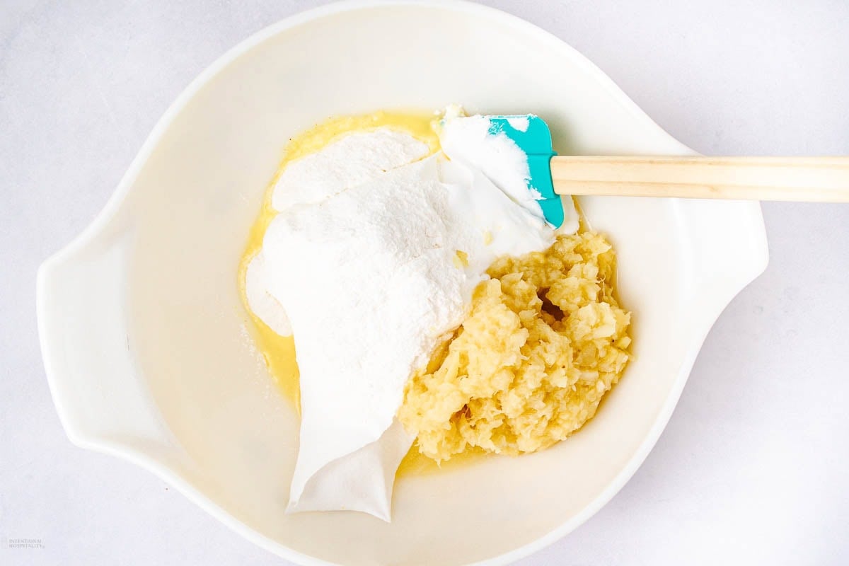 A white mixing bowl contains crushed pineapple, flour, sugar, and a creamy white ingredient, with a teal spatula resting on the side. The ingredients are unmixed and arranged in separate piles.