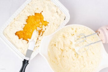 A baking dish with a partially spread creamy mixture surrounds a yellow jam-like filling, next to a bowl of whipped batter and a pink electric hand mixer on a white surface.