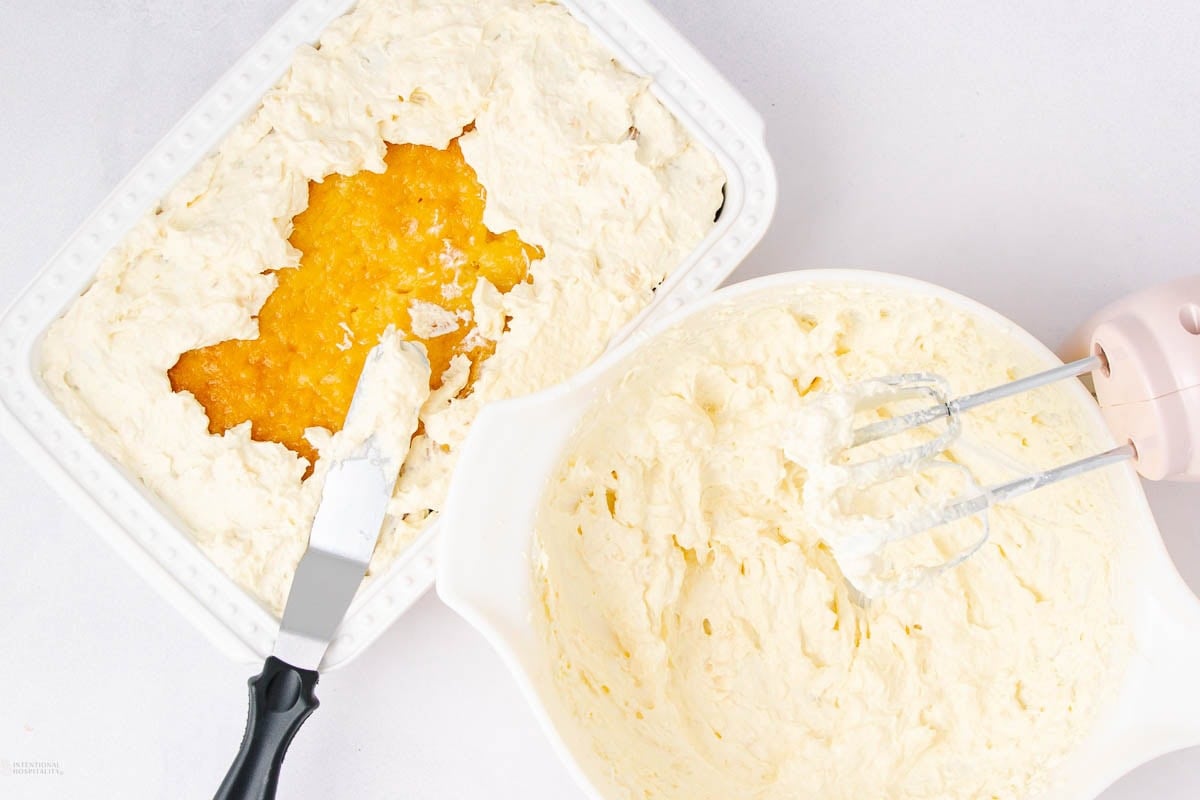 A baking dish with a partially spread creamy mixture surrounds a yellow jam-like filling, next to a bowl of whipped batter and a pink electric hand mixer on a white surface.