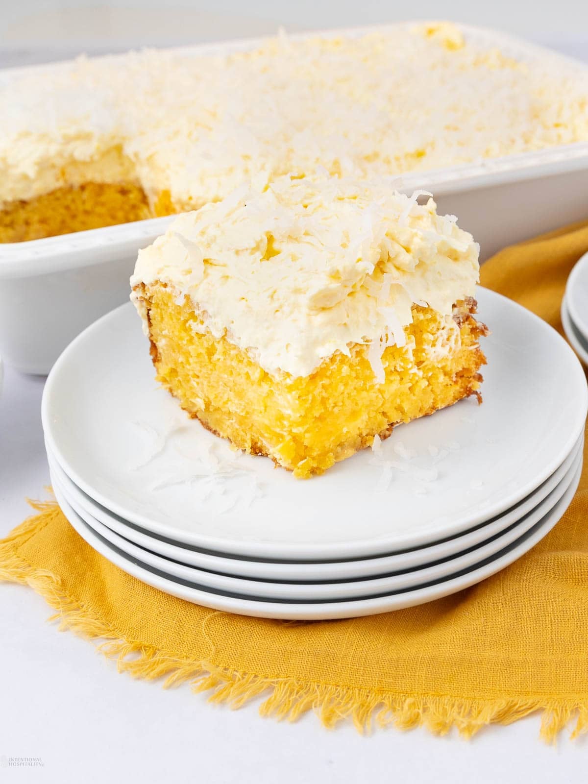 A slice of yellow poke cake with creamy frosting and shredded coconut on top sits on a stack of white plates, with the rest of the cake in a white baking dish in the background. The plates rest on a yellow cloth.