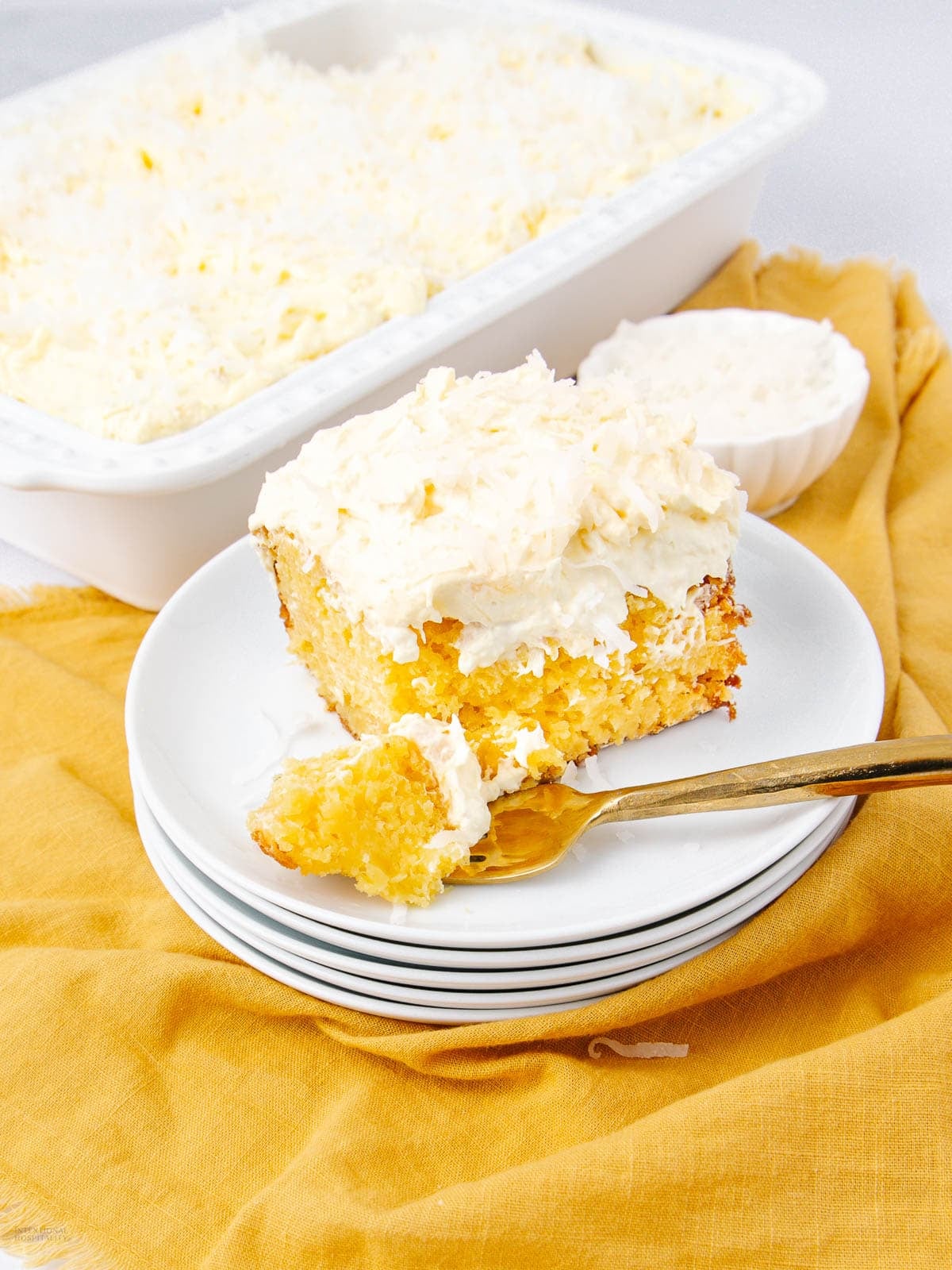 A plate with a slice of coconut cake topped with white frosting and shredded coconut, with a forkful taken out, sits on stacked white plates. In the background is a baking dish with more coconut cake.