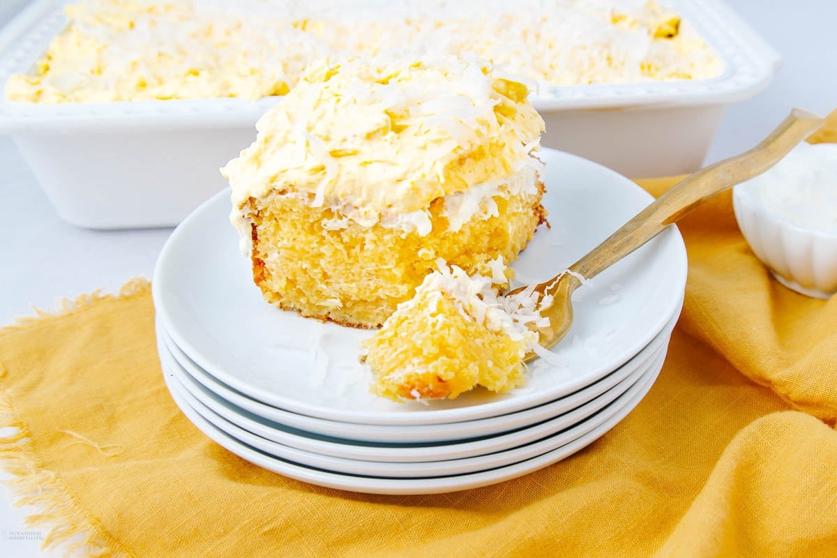 A slice of moist yellow cake topped with whipped cream and shredded coconut sits on a stack of white plates with a wooden fork. A baking dish and a bowl of coconut are in the background on a yellow cloth.
