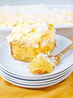 A slice of yellow coconut cake with white frosting and shredded coconut on top sits on a stack of white plates, with a fork holding a bite. A baking dish of the same cake is in the background on a yellow cloth.