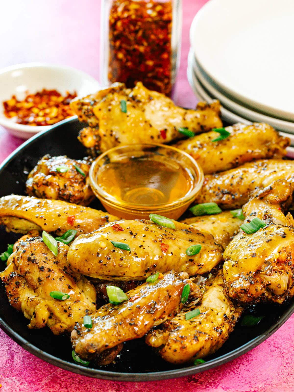 A black plate filled with seasoned chicken wings garnished with chopped green onions, served with a small bowl of dipping sauce. A bowl of red pepper flakes and a jar of seasoning are in the background.