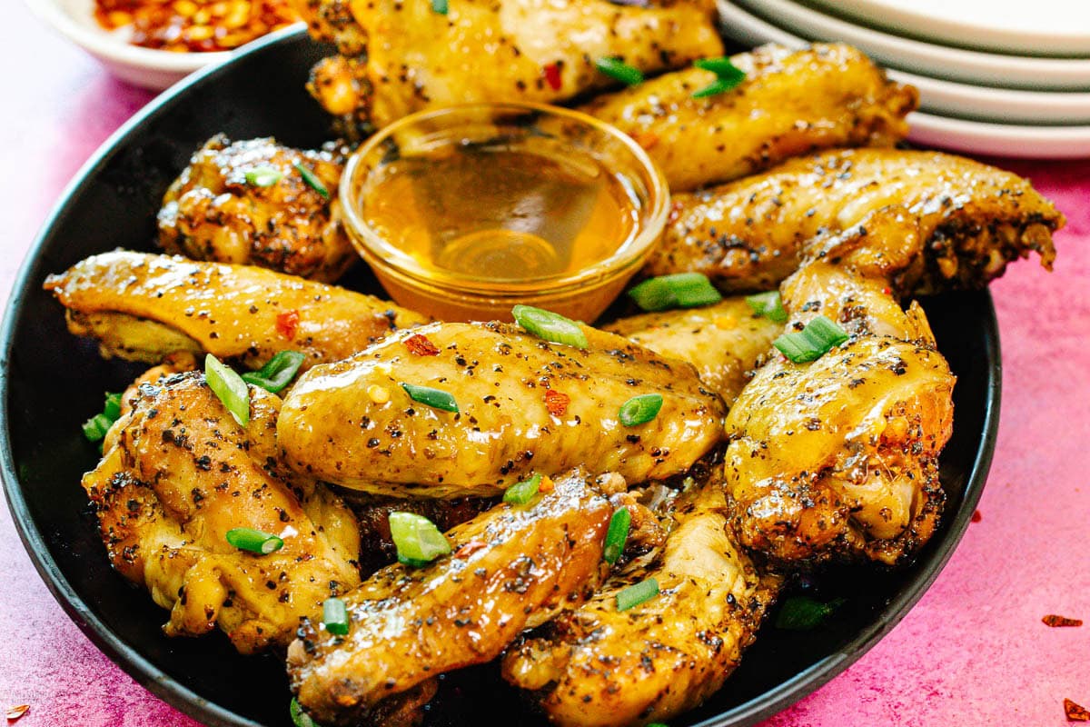 A black plate filled with seasoned chicken wings garnished with chopped green onions, served with a small cup of dipping sauce in the center; the background features stacked white plates and a pink surface.
