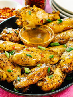 A plate of baked chicken wings seasoned with pepper and garnished with chopped green onions, served with a small bowl of honey dipping sauce. Red pepper flakes and stacked white plates are in the background.