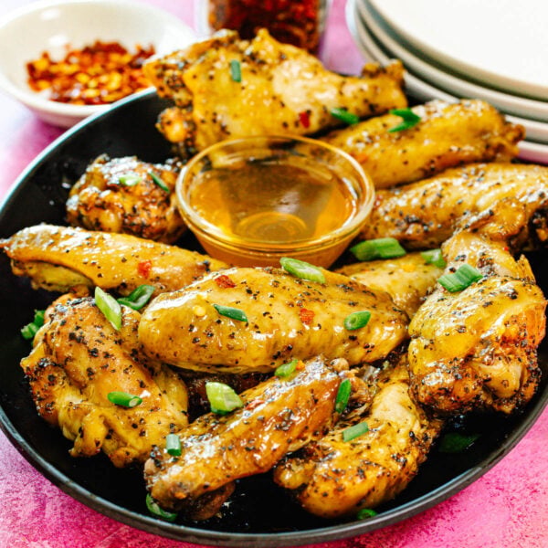 A plate of baked chicken wings seasoned with pepper and garnished with chopped green onions, served with a small bowl of honey dipping sauce. Red pepper flakes and stacked white plates are in the background.