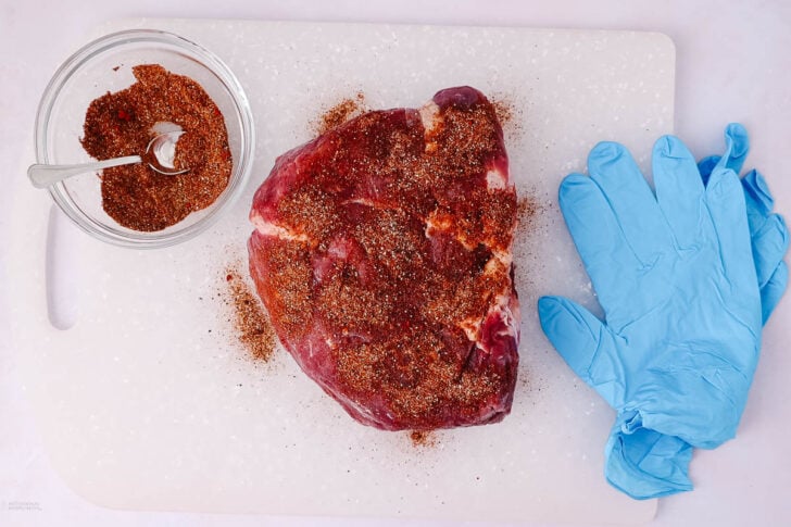 A raw piece of seasoned meat sits on a white cutting board next to a bowl of spice mix with a spoon, and a pair of blue disposable gloves.