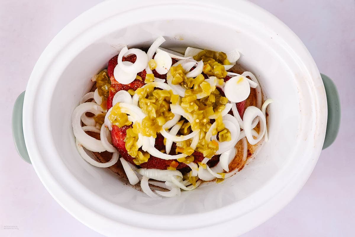 A slow cooker filled with sliced onions, chopped green chilies, and tomatoes on top of raw meat, ready to be cooked.