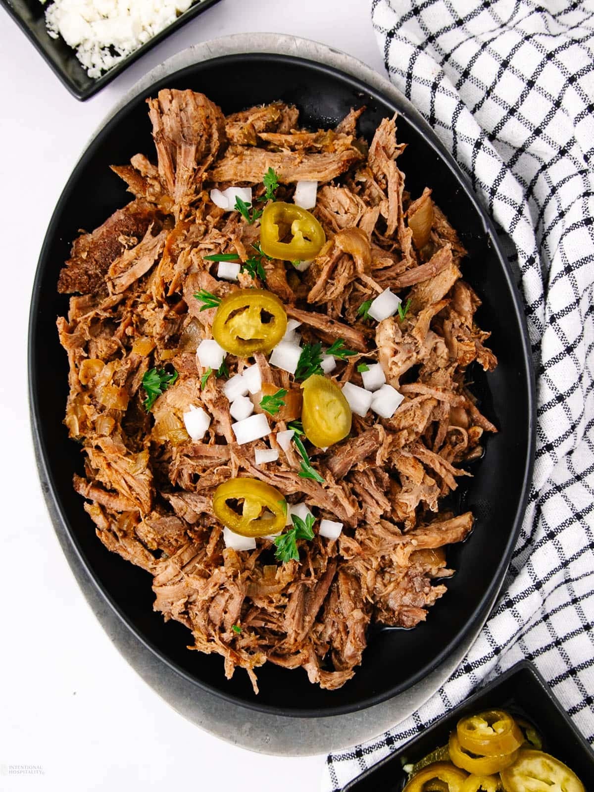 A black oval plate filled with shredded pork garnished with chopped onions, jalapeño slices, and parsley, placed next to a black-and-white checkered cloth and bowls of rice and other sides.