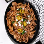 Shredded beef topped with chopped onions, sliced jalapeños, and fresh parsley, served on a black oval plate next to a white kitchen towel with black stripes.