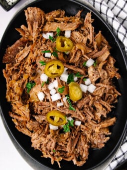 Shredded beef topped with chopped onions, sliced jalapeños, and fresh parsley, served on a black oval plate next to a white kitchen towel with black stripes.