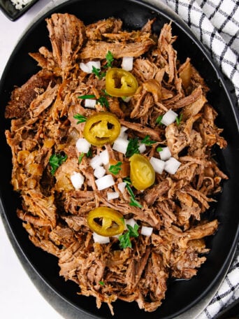 Shredded beef topped with chopped onions, sliced jalapeños, and fresh parsley, served on a black oval plate next to a white kitchen towel with black stripes.