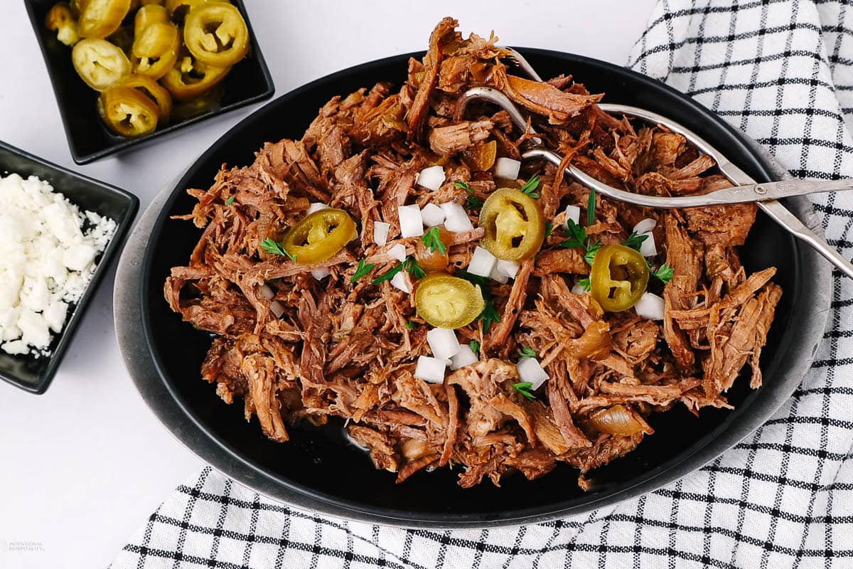 A black plate filled with shredded beef topped with diced onions, jalapeño slices, and chopped herbs. Nearby are small bowls of jalapeños and crumbled cheese, with a checkered cloth underneath.