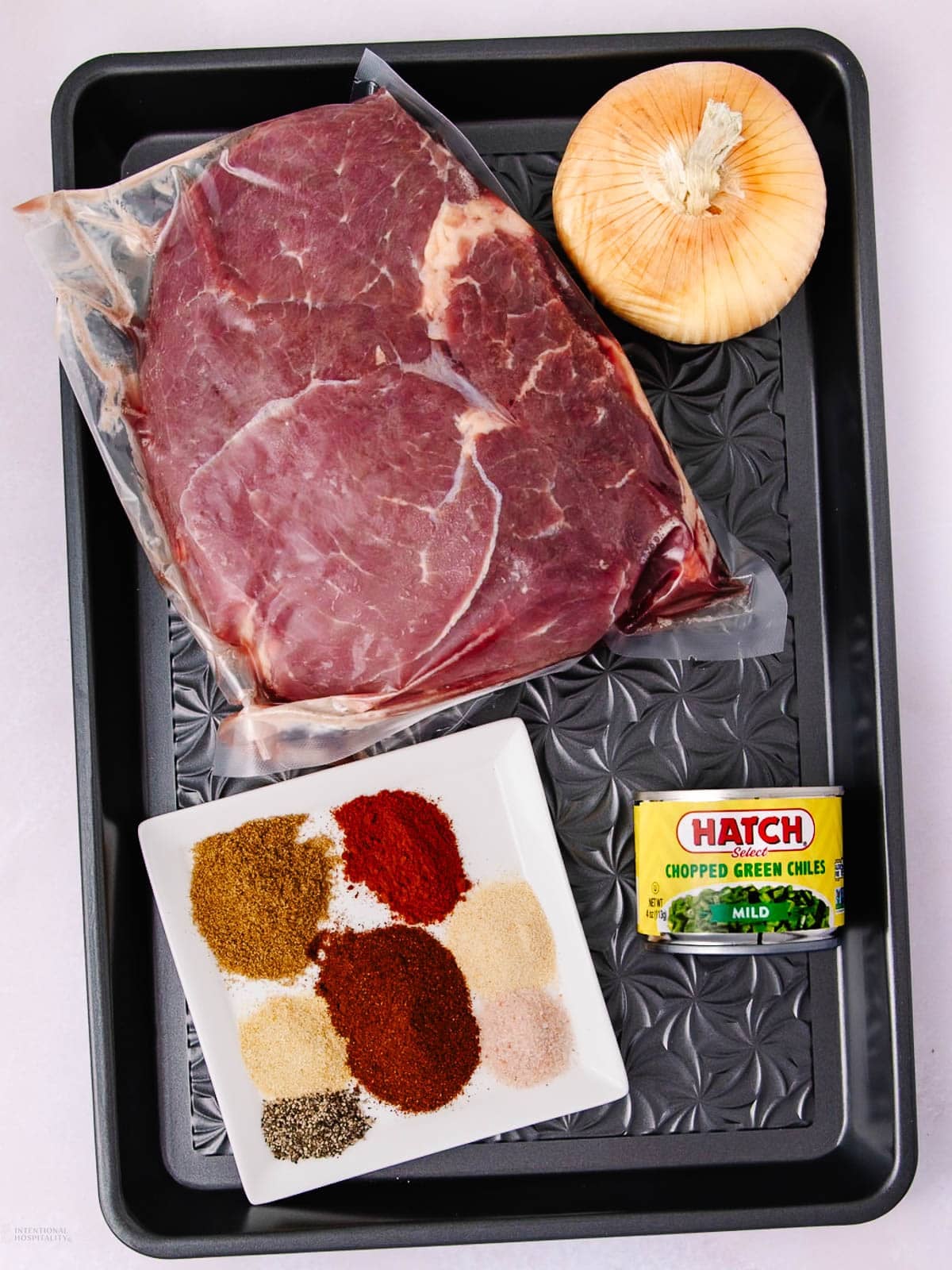 A tray with a large cut of raw beef in plastic wrap, a whole yellow onion, a can of mild chopped green chiles, and a square plate holding piles of various ground spices.