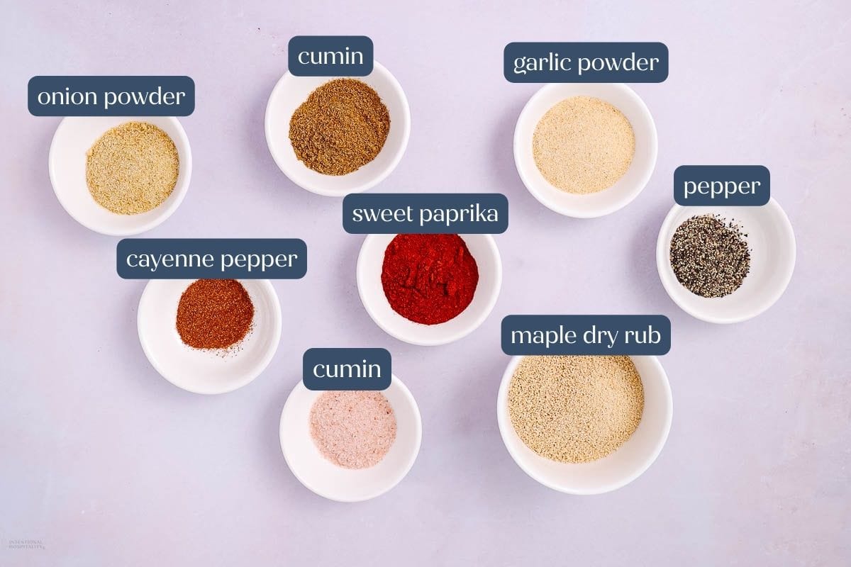 Eight small bowls containing different spices are arranged on a light surface. Each bowl is labeled: onion powder, cumin (two bowls), garlic powder, pepper, cayenne pepper, sweet paprika, and maple dry rub.