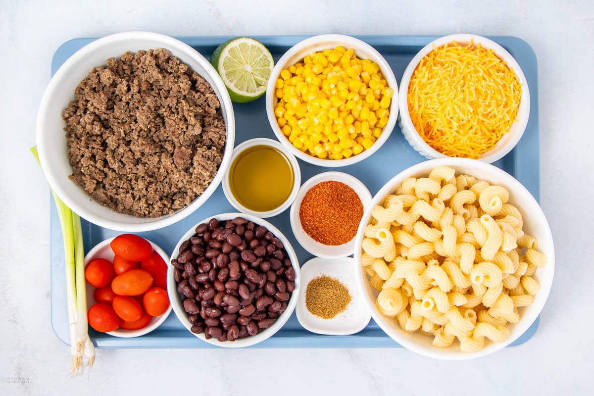 A tray with bowls of cooked ground beef, corn, shredded cheese, cooked pasta, black beans, grape tomatoes, green onions, lime half, oil, taco seasoning, and another spice on a white surface.