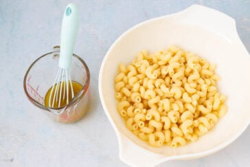 A white bowl filled with cooked spiral pasta sits next to a measuring cup containing a whisk and some brown liquid on a light blue surface.