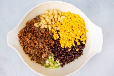 A large white bowl containing cooked ground meat, cooked pasta, yellow corn, black beans, and chopped green onions, arranged in separate sections on a light surface.