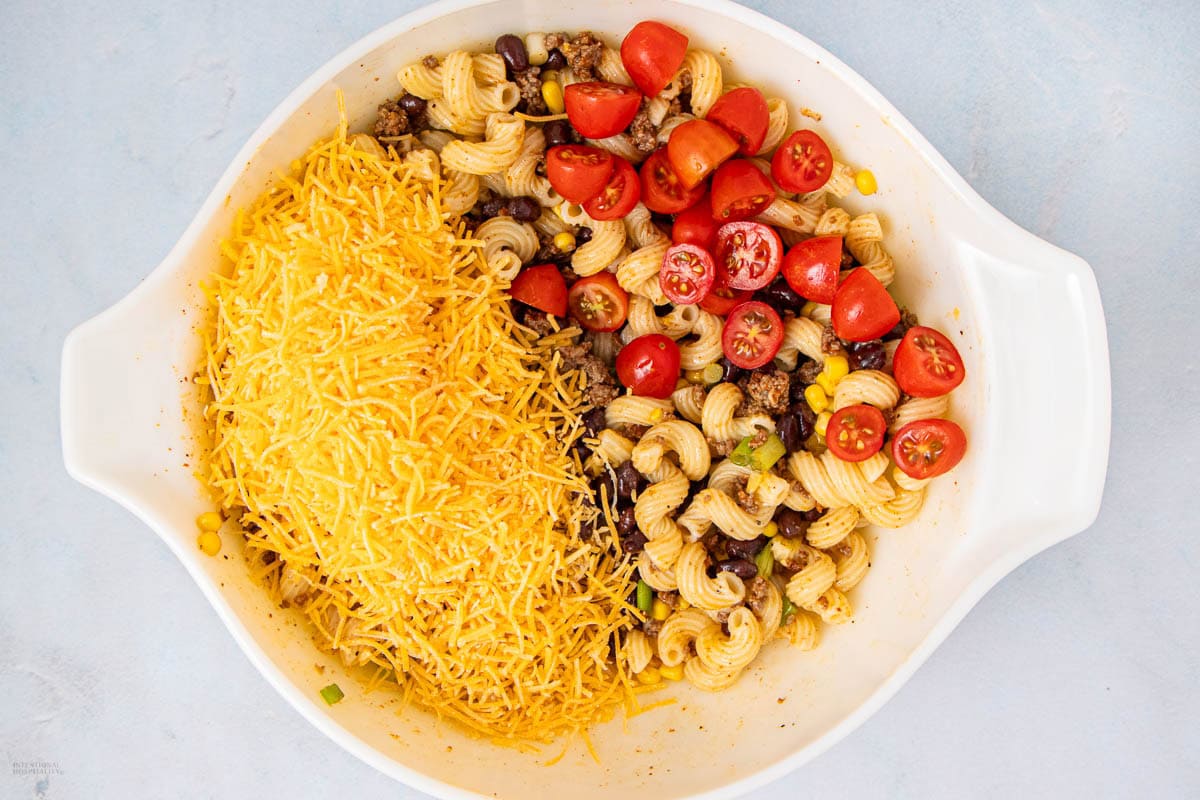 A white oval dish contains uncooked pasta salad ingredients: shredded cheddar cheese, spiral pasta, halved cherry tomatoes, black beans, corn, and ground beef, all separated and not yet mixed together.