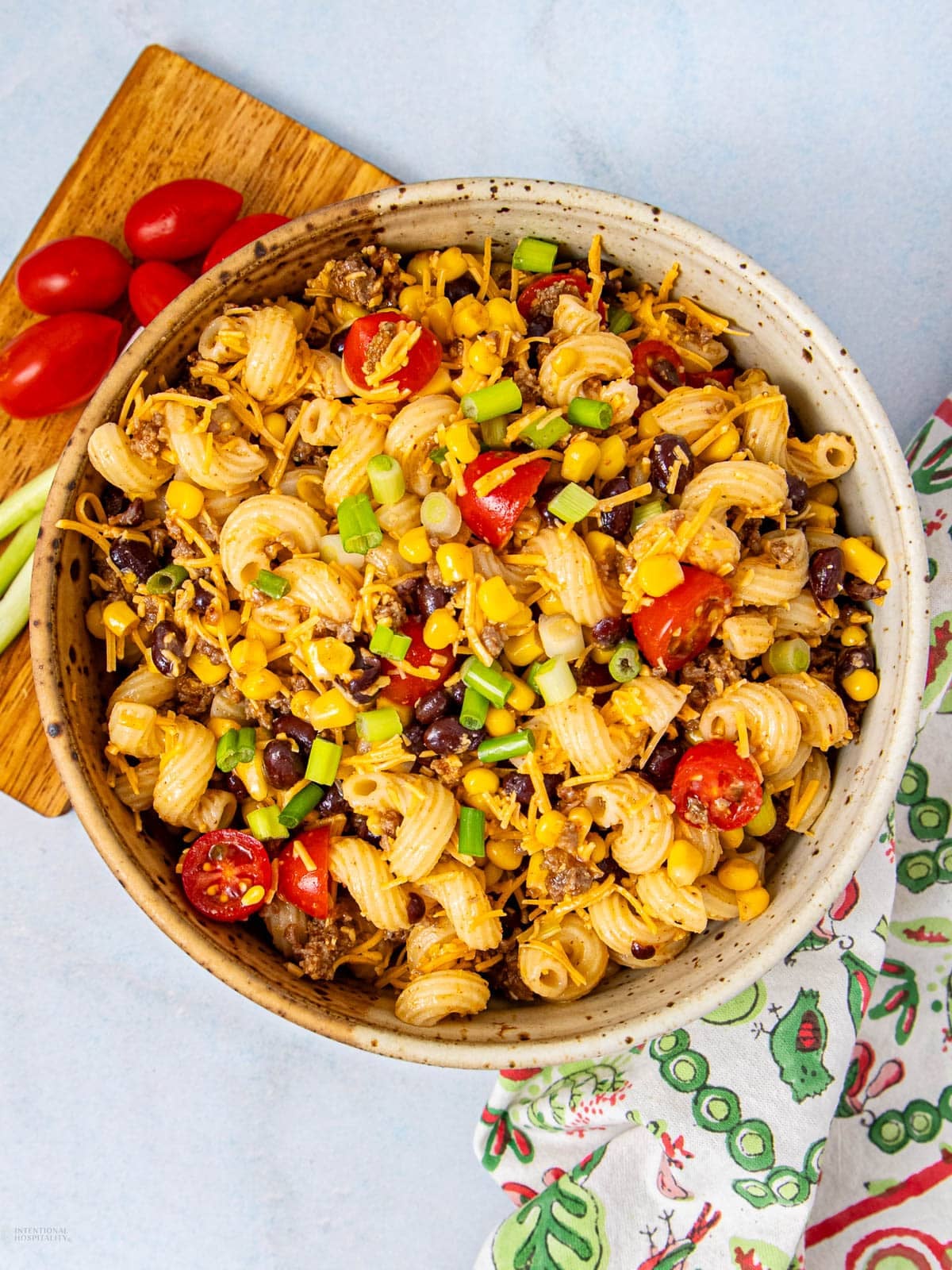 A bowl of pasta salad with cherry tomatoes, black beans, corn, ground meat, shredded cheese, and chopped green onions. A cutting board with grape tomatoes and scallions sits nearby, with a patterned napkin beside the bowl.