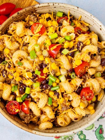 A bowl of pasta salad with spiral noodles, black beans, corn, cherry tomatoes, ground beef, shredded cheese, and chopped green onions. Some grape tomatoes are on a cutting board nearby, and a colorful napkin is in view.