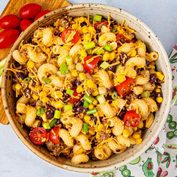 A bowl of pasta salad with spiral noodles, black beans, corn, cherry tomatoes, ground beef, shredded cheese, and chopped green onions. Some grape tomatoes are on a cutting board nearby, and a colorful napkin is in view.