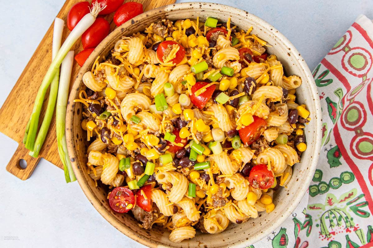 A bowl of pasta salad with spiral pasta, cherry tomatoes, black beans, corn, shredded cheese, and chopped green onions. Nearby are green onions, sliced tomatoes, and a colorful napkin.