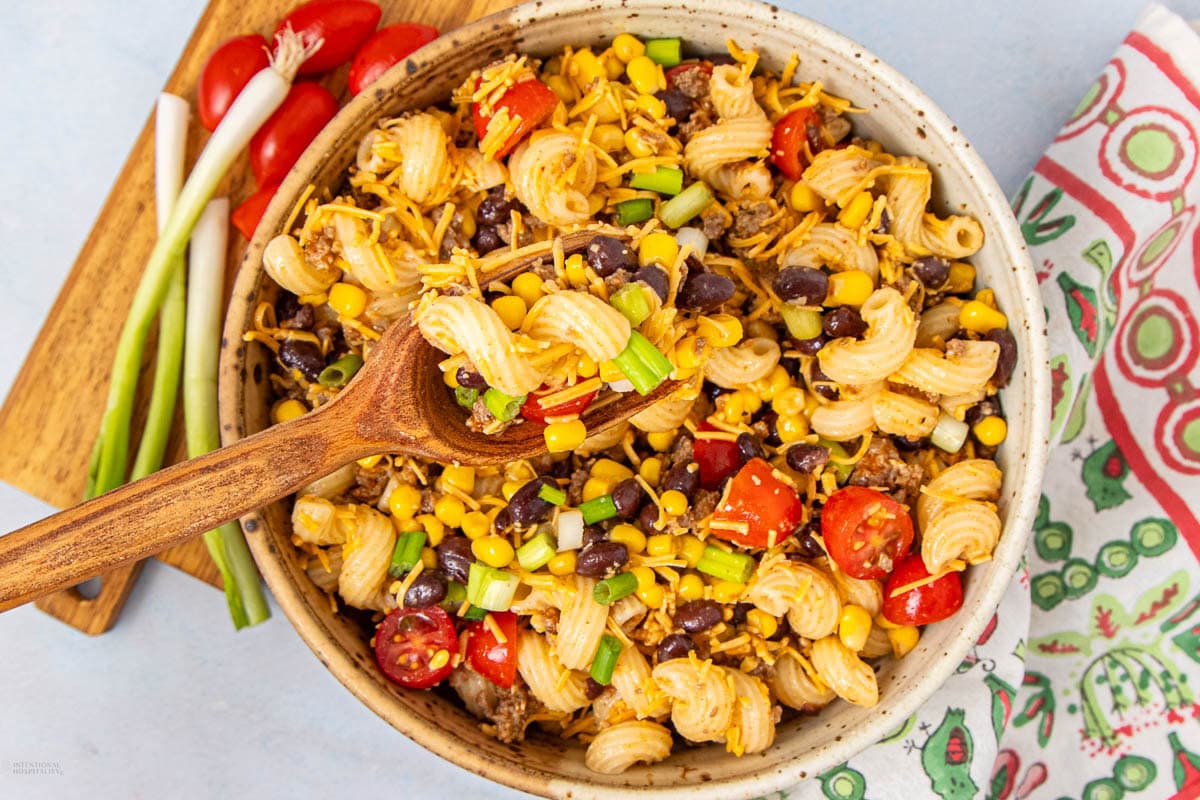 A bowl of pasta salad with spiral noodles, black beans, corn, cherry tomatoes, shredded cheese, and chopped green onions. A wooden spoon rests in the bowl, and green onions and tomatoes are on a wooden board nearby.