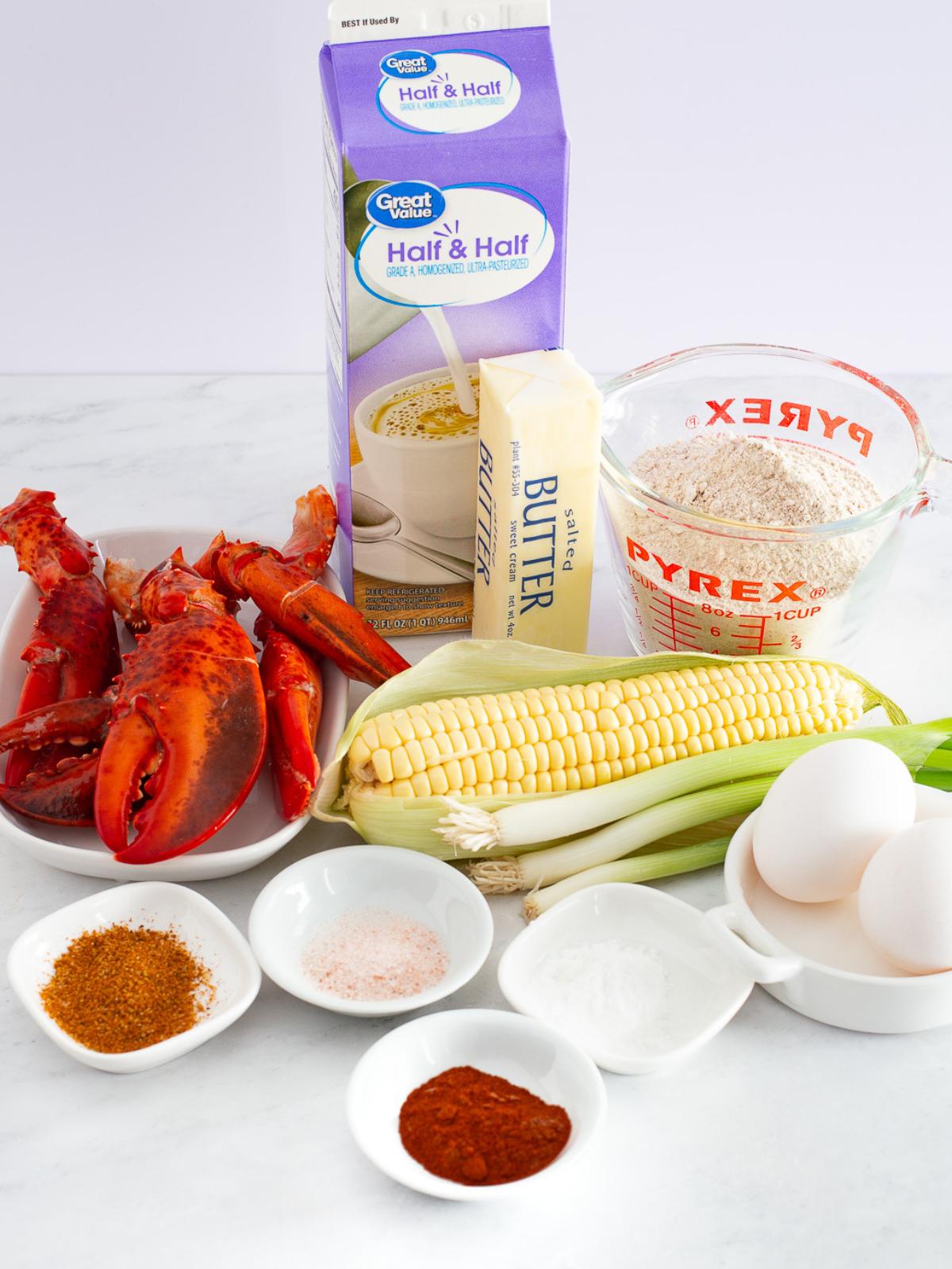 A variety of ingredients on a white surface: lobster claws, a stick of butter, half and half, a measuring cup of flour, an ear of corn, green onions, two eggs, and small bowls of spices and seasoning.