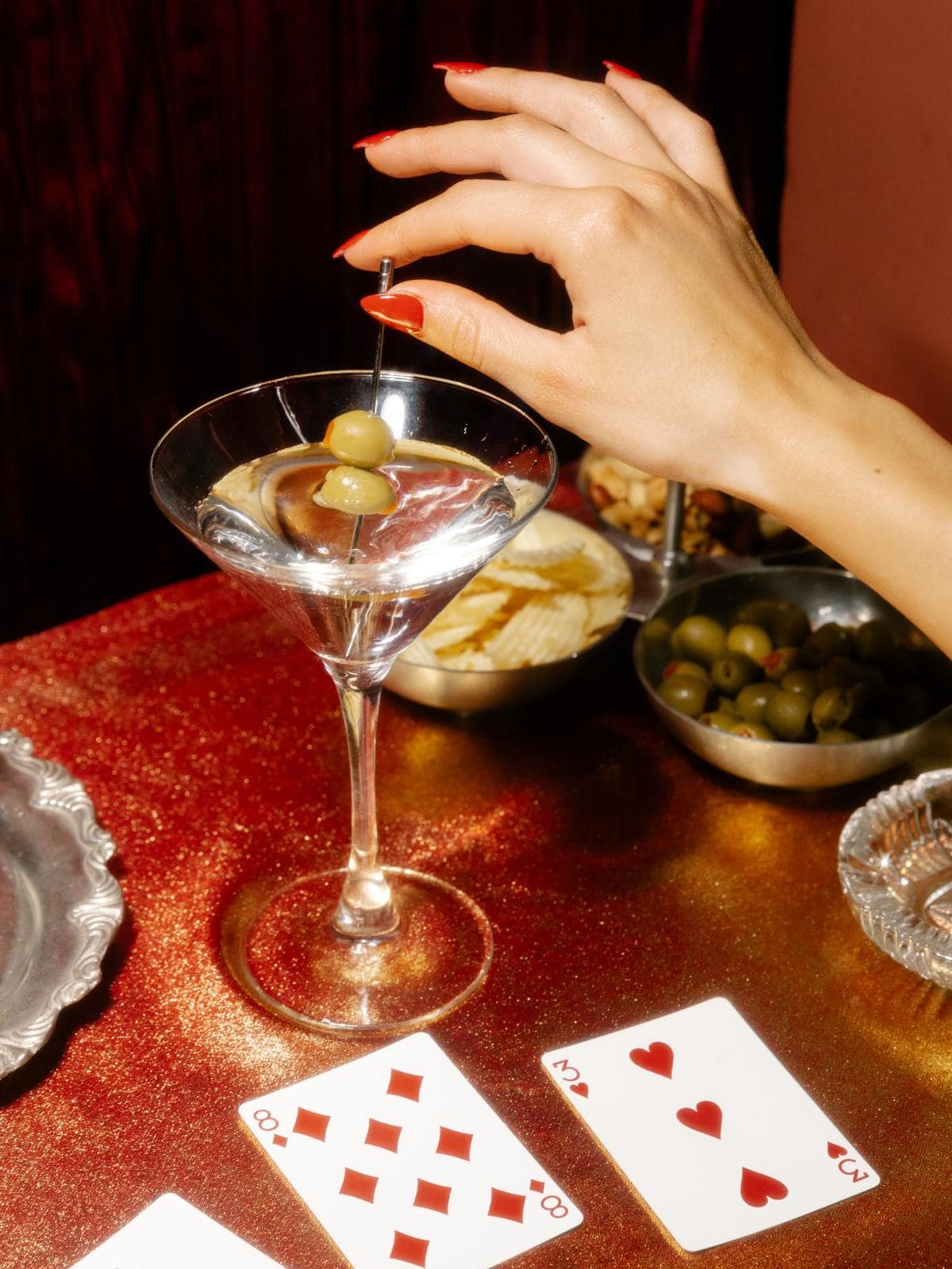 A hand with red nail polish holds a cocktail pick with olives over a martini glass on a glittery surface. Snack bowls and playing cards, including the eight of diamonds and two of hearts, are on the table.
