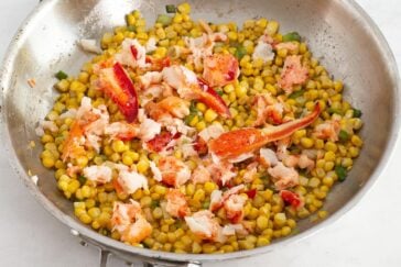 A stainless steel skillet filled with cooked corn kernels, green herbs, and large chunks of lobster meat on a light surface.