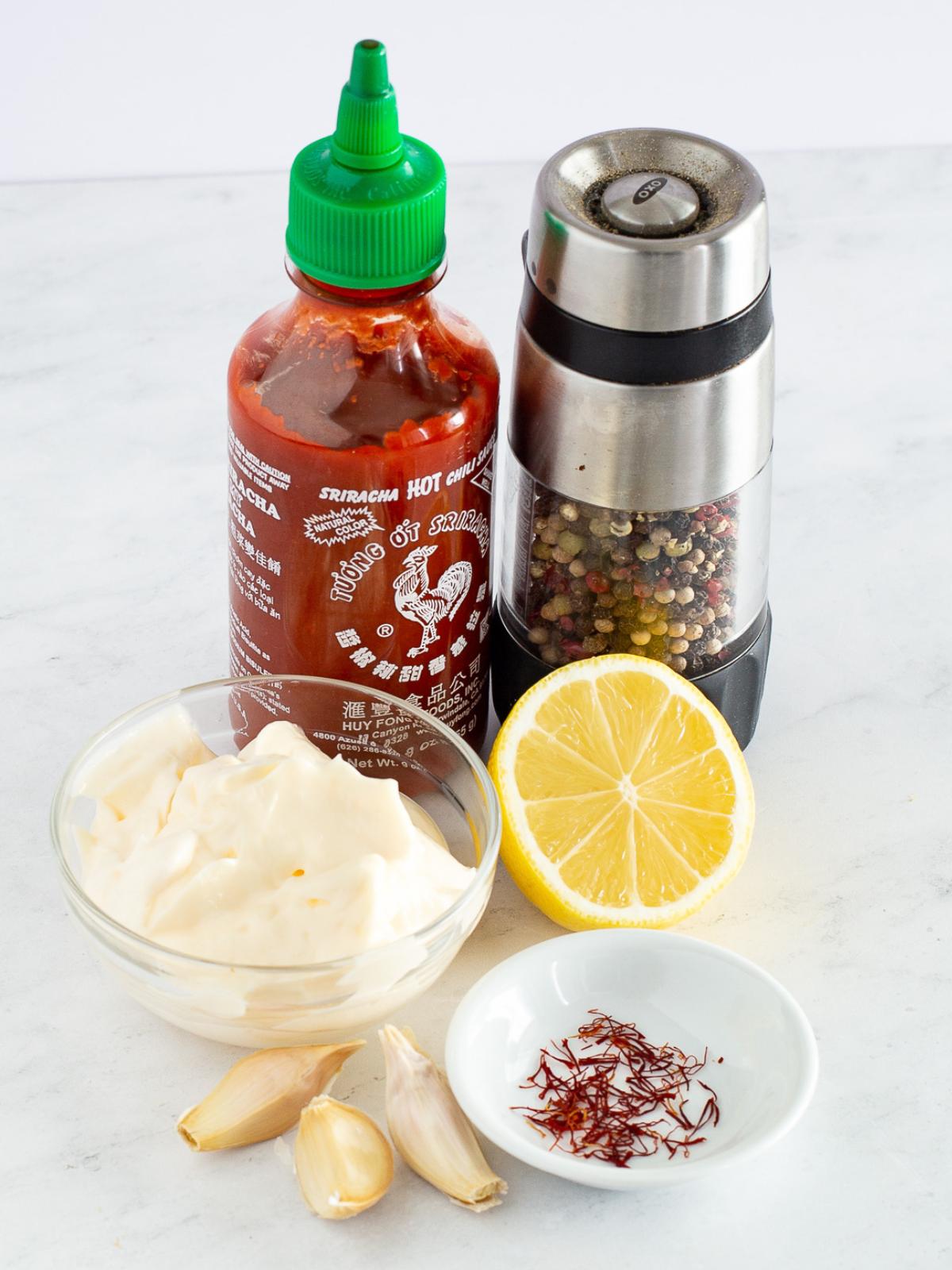 A bottle of sriracha, a pepper grinder, a halved lemon, a small bowl of mayonnaise, three garlic cloves, and a dish of saffron threads on a white surface.