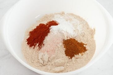 A white bowl contains flour with separate piles of red spices, pink salt, white baking powder, and brown seasoning on top, ready to be mixed.