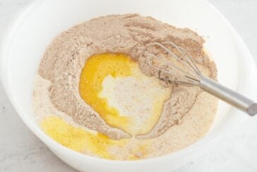 A mixing bowl containing flour, cocoa powder, melted butter, and milk with a whisk partially submerged, ready for mixing.