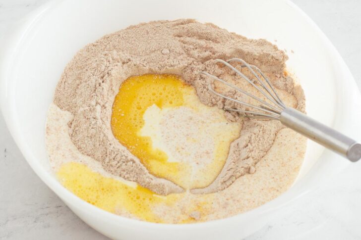 A mixing bowl containing flour, cocoa powder, melted butter, and milk with a whisk partially submerged, ready for mixing.