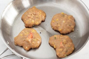 Four uncooked vegetable fritters with visible corn and chopped peppers in a metal frying pan, ready to be cooked.