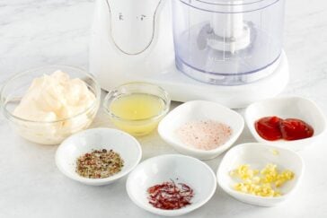 A food processor sits behind small bowls containing mayonnaise, lemon juice, pink salt, ketchup, minced garlic, ground pepper, and saffron threads on a white countertop.
