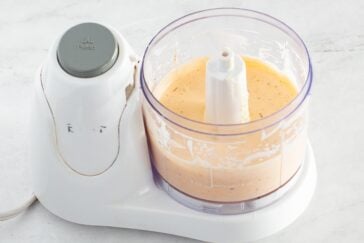 A small food processor filled with a creamy, light orange sauce sits on a white countertop. The lid is off, revealing the smooth mixture inside the clear container.