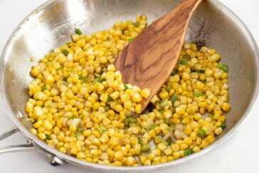 Corn kernels being sautéed with chopped green onions in a stainless steel pan, stirred with a wooden spatula.