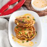 Two golden brown corn fritters topped with chopped green onions and lemon zest, served on a white plate with a creamy dipping sauce. Red tongs and a bowl of extra sauce are nearby on a wooden board.