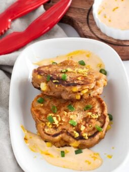 Two golden brown corn fritters topped with chopped green onions and lemon zest, served on a white plate with a creamy dipping sauce. Red tongs and a bowl of extra sauce are nearby on a wooden board.